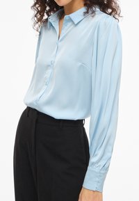 Femme portant un chemisier en satin bleu clair à manches longues, rentré dans un pantalon noir taille haute, visible du cou aux hanches.