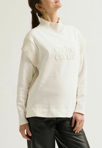 Crèmekleurige sweatshirt met ribbelaccenten op de schouders en een hoge kraag, met reliëftekst die "cache coeur" zegt. Zachte stof textuur.
