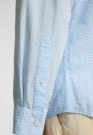 Chemise bleu clair en tissu texturé, avec des poignets à bouton et de petits boutons. Le design présente un motif subtil et une forme de poignet pointue.