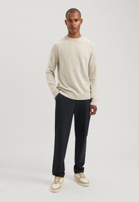 Beige gebreide trui met een ronde hals, gecombineerd met donkere broeken. De schoenen zijn crèmekleurige en beige sneakers. Eenvoudig ontwerp, zachte textuur.