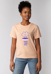Perzikkleurig katoenen t-shirt met een blauwe grafische afbeelding van een lachende plant en de tekst "GOEDE VIBES ALLEEN" eronder, ronde halslijn, korte mouwen.