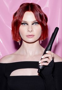 Une personne avec des cheveux courts roux et un maquillage des yeux noir audacieux tient un outil de coiffure GHD Mini noir contre un fond rose.