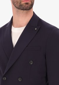 Blazer doppiopetto blu navy in tessuto liscio con bottoni neri, reverse a punte e taschino sul petto. Indossato sopra una camicia bianca.