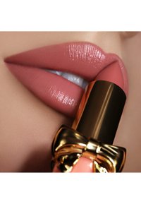 Matter nuder Lippenstift wird auf volle Lippen aufgetragen. Der Lippenstiftbehälter ist mit einer goldenen Schleife verziert und hat ein glänzendes Metallfinish.