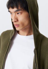 Oliva zöld cipzáros kapucnis pulóver, texturált kötött anyagból, kapucnival, fehér crewneck póló fölött viselve. Diszkrét, minimalista dizájn.