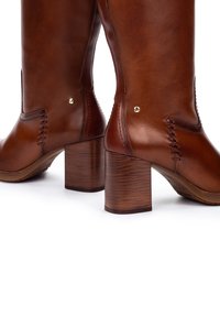 Bottes en cuir marron jusqu'aux mollets avec des talons en bois épais et des coutures décoratives sur un fond blanc.