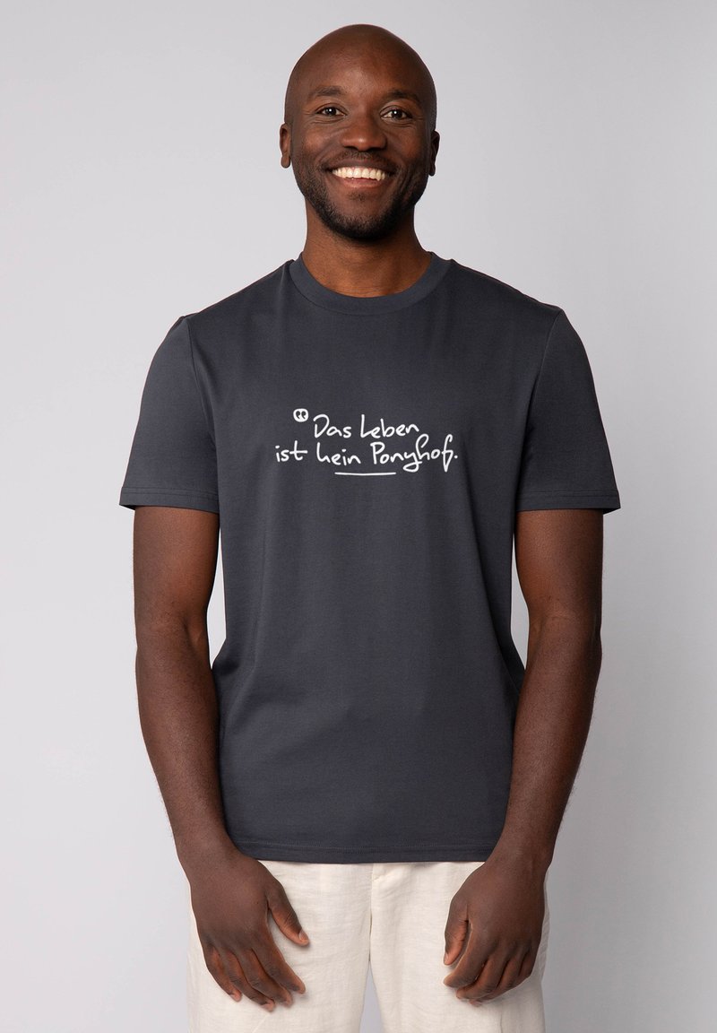 Lächelnder Mann, der ein dunkelgraues T-Shirt mit handgeschriebenem deutschen Text "Das Leben ist kein Ponyhof" und hellbeige Hosen trägt, vor einem einfarbigen Hintergrund steht.