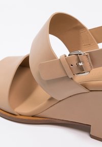 Sandale compensée à talon haut beige avec de larges brides croisées, texture en cuir lisse et une bride de cheville ajustable avec détail de boucle en argent.