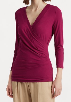 Femme portant un haut cache-cœur à manches longues de couleur rose profond associé à un pantalon beige ample, sur un fond uni.