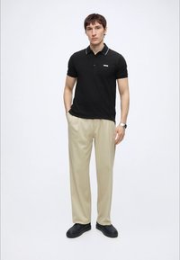 Ung mand stående med hænderne i lommerne, iført sort poloshirt, beige bukser, sorte sko og et armbåndsur på hvid baggrund.