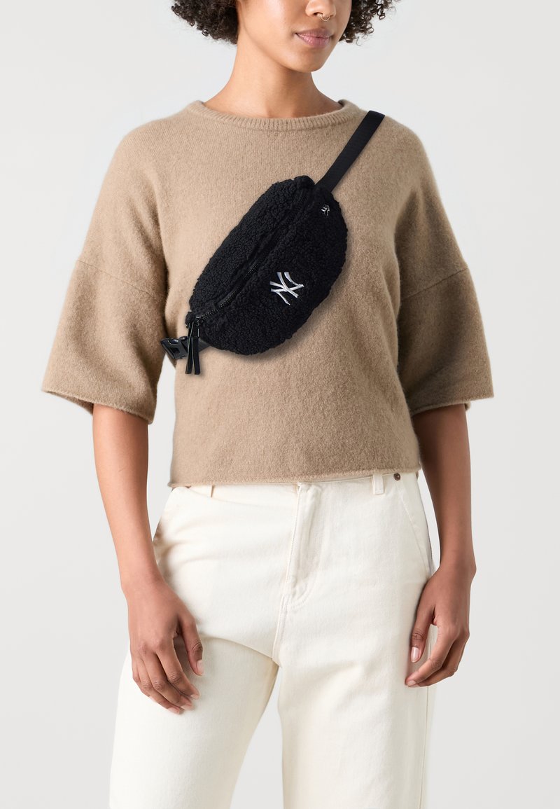 Svart fleece crossbody-väska med dragkedja och logotypdetalj. Bärs över en beige tröja och matchas med ljusa byxor.