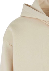 Felpa beige con un tessuto morbido e strutturato. Presenta un cappuccio con coulisse e spalle ripiegate per una vestibilità rilassata.