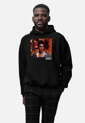 Black hoodie featuring a front graphic of a man against a colorful sunset background. Text "SCARFACE" in white below the image.