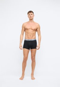Černé bavlněné boxerky s bílým páskem s logem Nike; přiléhavý design; modelka stojící na boso na jednobarevném pozadí.