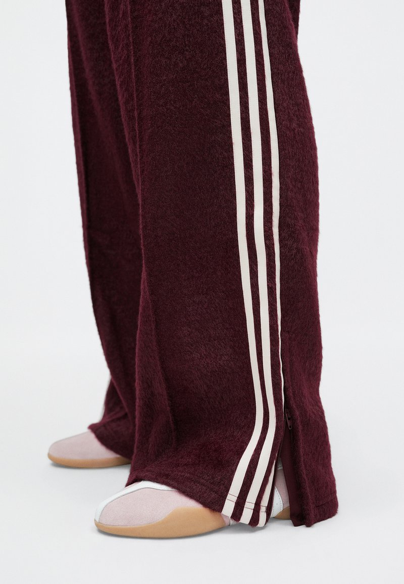 Pantaloni in fleece bordeaux con strisce laterali bianche e orli con zip, abbinati a scarpe rosa chiaro e beige con punta tondeggiante.