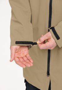 Beige wasserdichte Jacke mit hohem Kragen, schwarzem Reißverschluss und Klettverschluss zur Anpassung der Ärmel. Glatte Textur und leichtes Design mit einer lockeren Passform.