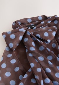 Braune, durchsichtige Bluse mit großen blauen Polka-Dots, mit einer gebundenen Schleife am Kragen und einem sichtbaren Next-Kleideretikett.