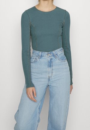 Mujer con una camiseta de manga larga ajustada en color teal con detalles de costuras visibles, combinada con unos vaqueros de pierna ancha de talle alto en azul claro.