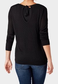 Frau trägt ein schwarzes, strukturiertes Langarmoberteil mit einem Schlüsselloch-Ausschnitt und einer gebundenen Schleife am Rücken, kombiniert mit blauen Jeans.