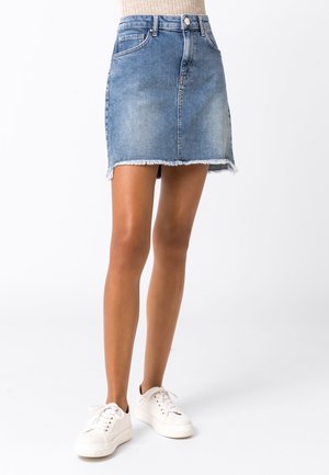 Mujer con una minifalda de mezclilla azul claro con borde deshilachado y zapatillas blancas, posando frente a un fondo blanco liso.