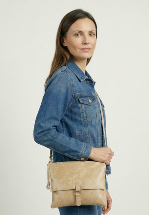 Kvinde i denimjakke, der bærer en beige lædertaske med spænde mod en ensartet lys baggrund.