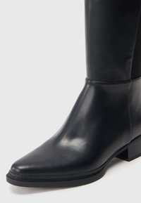 Botte en cuir noir mi-mollet avec bout pointu, petit talon bloc, et finition lisse sur un fond clair.