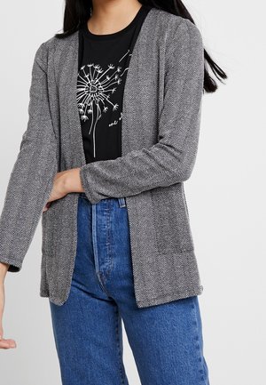 Vrouw draagt een grijze visgraatblazer over een zwart t-shirt met een witte paardenbloemprint en blauwe jeans met hoge taille tegen een witte achtergrond.