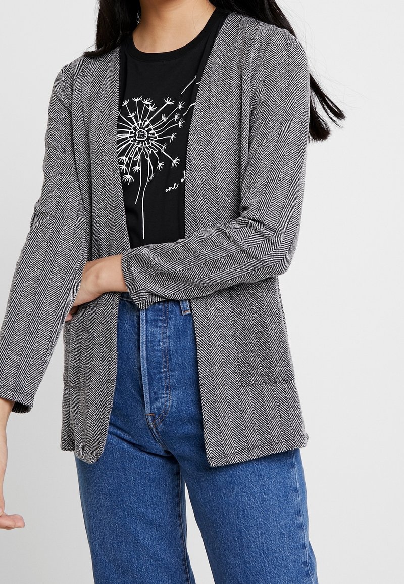 Femme portant un blazer en chevron gris sur un t-shirt noir avec un motif pissenlit blanc et un jean taille haute bleu sur fond blanc.