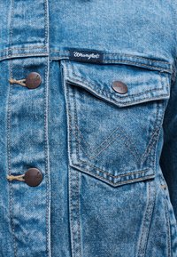 Κοντινή λήψη μπροστινής τσέπης ανοιχτό μπλε τζιν μπουφάν με καφέ μεταλλικά κουμπιά και ορατή ετικέτα της μάρκας Wrangler.