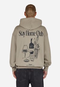 Khaki hettegenser med et svart grafisk design av drikkevarer og planter på baksiden, med ribbestrikkede mansjetter og kant.