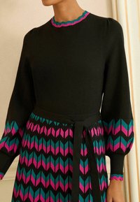 Hosszú ujjú ruha fekete felsőrésszel és teal és rózsaszín cikkcakk mintás szoknyával. Hullámos dekoltázzsal és megkötős derékkal rendelkezik.