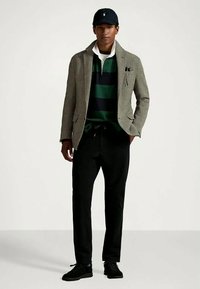 Homem com boné preto, blazer cinza, camiseta de rugby listrada em verde e preto, calças pretas e ténis pretos, de pé contra um fundo branco.