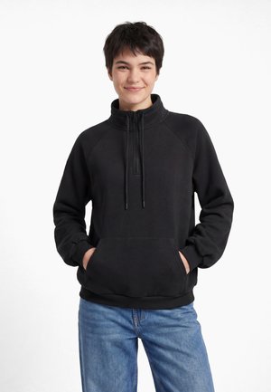 Giovane con capelli corti e scuri che sorride, indossa un pullover nero con mezza cerniera, con le mani nella tasca frontale e jeans blu.