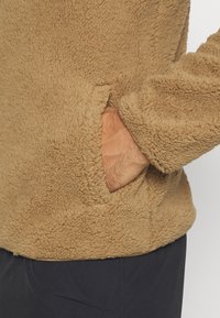 Pull-over en polaire marron avec une surface texturée, doté d'une poche latérale et d'un tissu doux, accompagné de pantalons sombres.
