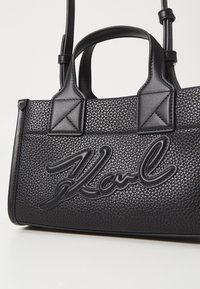 Černá texturovaná kabelka s dvěma krátkými ušima, s embosovaným logem. Materiál má jemně zrnitý povrch, který přidává hloubku designu.