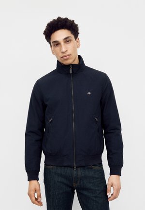 Ung mand med krøllet hår iført en mørk navy lynlås jakke og mørke jeans, stående mod en ensfarvet lys baggrund.