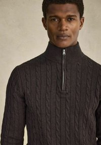 Mörkgrå stickad tröja med hög krage och dragkedja. Har en kabeltextur och ribbad detaljering längs halsen och ärmarna.