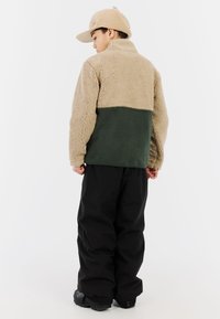 Tan en groene fleecedonsjack met hoge kraag, van achteraf bekeken, gecombineerd met zwarte broeken en een tan kleurige pet. Zachte textuur en kleurblokontwerp.