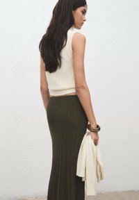 Top de tricot fără mâneci crem, fustă olive în dungi, și o bluză ușoară ținută în mână. Păr lung și drept. Fundal simplu și neted.