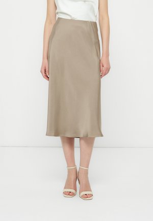 Kobieta w beżowej satynowej spódnicy midi, białym topie bez rękawów i beżowych sandałach na obcasie z paskiem wokół kostki, stojąca na białym tle.