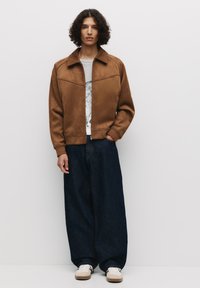 Veste bomber en suede marron avec fermeture éclair à l'avant, poignets et col côtelés, associée à un jean en denim foncé à jambes larges et des baskets beiges.