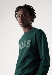 Felpa verde con maniche lunghe, caratterizzata da un grande logo "BOSS" grigio chiaro sul davanti. Realizzata in un morbido tessuto testurizzato.