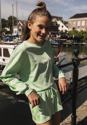 Jong meisje in een lichtgroene trui en bijpassende rok staat bij een zwarte reling vlakbij het water met boten en gebouwen op de achtergrond.