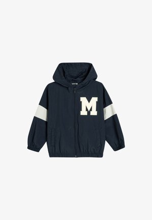 Marineblauwe jas met capuchon, elastische manchetten en een "M"-logo. Heeft witte gestreepte accenten op de mouwen. Gemaakt van lichte stof.
