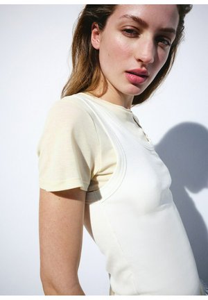 Kvinde med lyst brunt hår iført en creme kortærmet skjorte og en ærmeløs hvid top, stående mod en hvid væg med skygge.