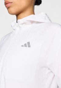 Geacă albă ușoară, cu fermoar și glugă, având un logo argintiu pe partea stângă a pieptului și o textură netedă, mată.