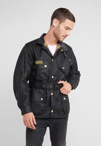 Black waxed jacket featuring a belted waist, golden buttons, four front pockets, and a corduroy collar with a plaid lining detail.
