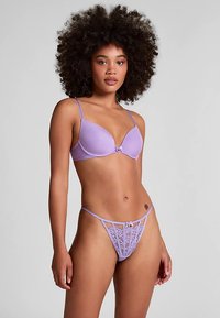 Ensemble soutien-gorge et string en tissu lisse couleur lavande. Le soutien-gorge possède des bonnets moulés et des bretelles réglables ; le string comporte un devant en dentelle et des fines bretelles latérales.