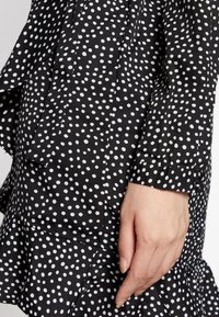 Svart skjorta med vita prickar, med en volangkant och ärmar med manschett. Tyget verkar vara slätt med en lätt lyster.