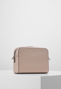 Suuri beige ristiselkäreppu, jossa on teksturoitu pinta ja hopea vetoketju, asetettuna valkoiselle pedestaalille tasaisella taustalla.